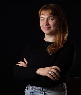 Das Bild zeigt eine FLINTA*-Person mit langen, roten Haaren und einem Pony. Sie verschränkt ihre Arme und lächelt.