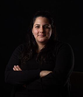 Das Bild zeigt eine FLINTA*-Person mit langen, dunkelbrauen Haaren. Sie verschränkt ihre Arme und lächelt.