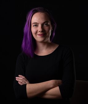 Das Bild zeigt eine FLINTA*-Person mit schulterlangen, lilanen Haaren. Sie verschränkt ihre Arme und lächelt.