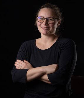 Das Bild zeigt eine FLINTA*-Person mit zusammengebundenen, braunen Haaren. Sie trägt eine Brille. Sie verschränkt ihre Arme und lächelt.